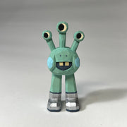 Mint green whittled wooden sculptures of a space alien, with 3 eyes and 2 legs without any other body features. It smiles with buck teeth and wears shiny silver shoes.
