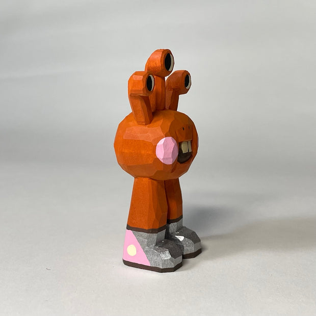 Orange red whittled wooden sculptures of a space alien, with 3 eyes and 2 legs without any other body features. It smiles with buck teeth and wears shiny silver shoes.