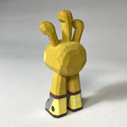 Yellow whittled wooden sculptures of a space alien, with 3 eyes and 2 legs without any other body features. It smiles with buck teeth and wears shiny silver shoes.