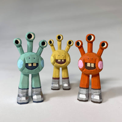3 painted whittled wooden sculptures of space aliens, with 3 eyes and 2 legs without any other body features. They all smile with buck teeth and wear shiny silver shoes.