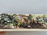 Diorama style mixed media sculpture in a long rectangular glass box of a forest floor scene, with 2 frogs dueling in the center. All around them are various cut paper leaves and plants.