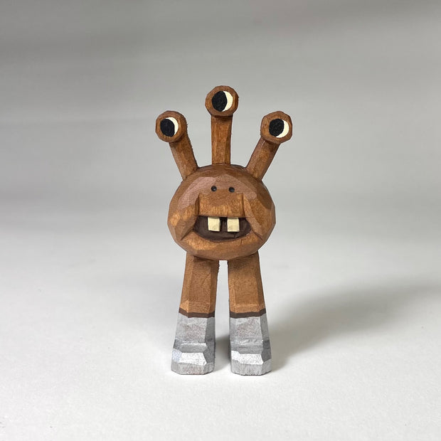 Natural wood whittled wooden sculptures of a space alien, with 3 eyes and 2 legs without any other body features. It smiles with buck teeth and wears shiny silver shoes.