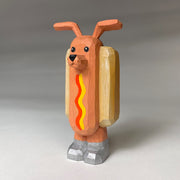 Whittled wooden sculpture of a hot dog, standing up and wearing silver shoes. It has a face of a dog and large protruding dog ears. It has no arms and a squiggle of mustard and ketchup on its front.
