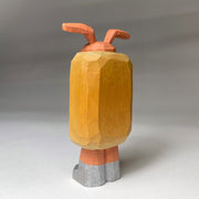 Whittled wooden sculpture of a hot dog, standing up and wearing silver shoes. It has a face of a dog and large protruding dog ears. It has no arms and a squiggle of mustard and ketchup on its front.