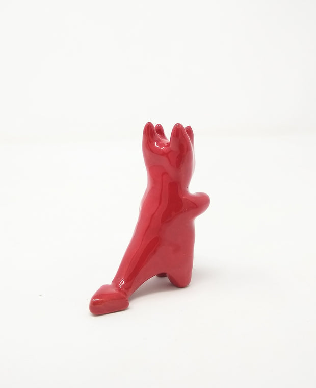 Red ceramic sculpture of a devil like creature, with a smooth body and simple smiling face. It has 4 horns atop its head and one arm extended out, as if waving.