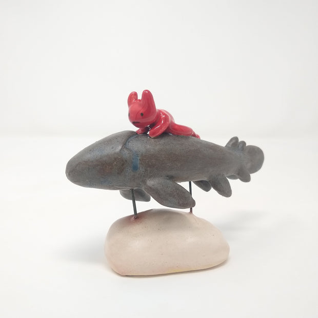 Ceramic sculpture of a grey fish with many fins and no facial features. A small red devil, with minimal facial features, rides atop of it. The pair are held up by 2 pieces of wire that lead to a cream colored mound that resembles stone.