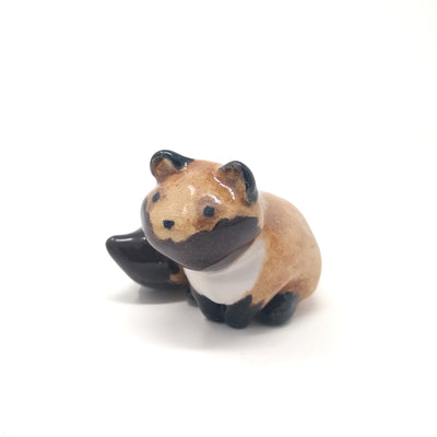 Small ceramic sculpture of a brown fox like creature, with small pointed ears and a large fluffy tail. It has painted on eyes and a simple dot nose.