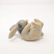 Off white ceramic sculpture with a crackled outer glaze of a rabbit, laying on its back with its feet up in the air and arms reaching its toes. Subtle blue coloring surrounds its neck and face.