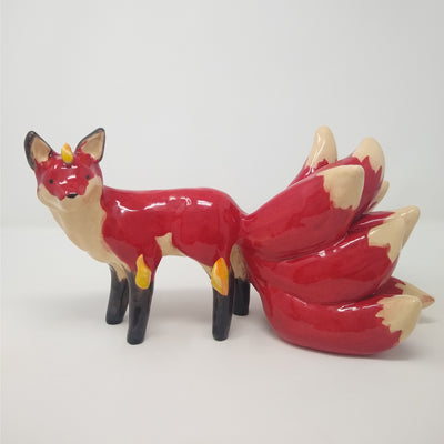Glossy ceramic sculpture of a red fox with a large tail, composed of 9 different smaller tails. The fox has a small golden horn and some gold flames come of its body.