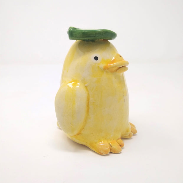 Ceramic sculpture of a yellow duck with a green leaf atop its head.