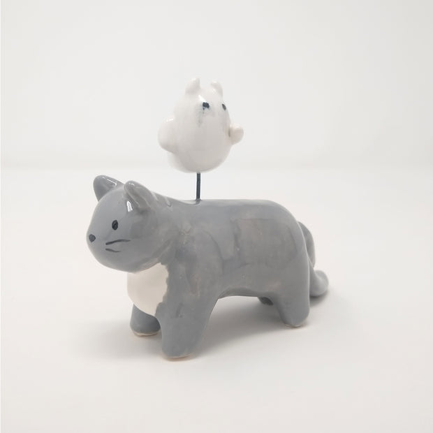 Ceramic sculpture of a gray cat with simplistic body features and a drawn on eyes and whiskers. Hovering atop its back is a small white ghost, with watercolor style tears.