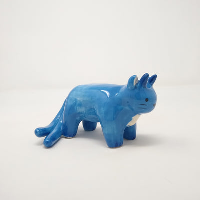 Small blue ceramic sculpture of a cat with 2 tails and horns coming out atop its head. It has simplistic body features and a drawn on face.