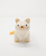 White ceramic cat with simplistic bodily features and a drawn on eyes and nose. Atop its ears, tail and feet is a subtle orange coloring.