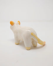 White ceramic cat with simplistic bodily features and a drawn on eyes and nose. Atop its ears, tail and feet is a subtle orange coloring.