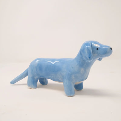 Small ceramic sculpture of a blue Dachshund, with a long tail and white underbelly and marks above its eyes, like raised eyebrows.