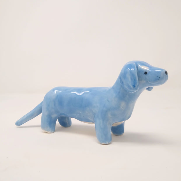 Small ceramic sculpture of a blue Dachshund, with a long tail and white underbelly and marks above its eyes, like raised eyebrows.