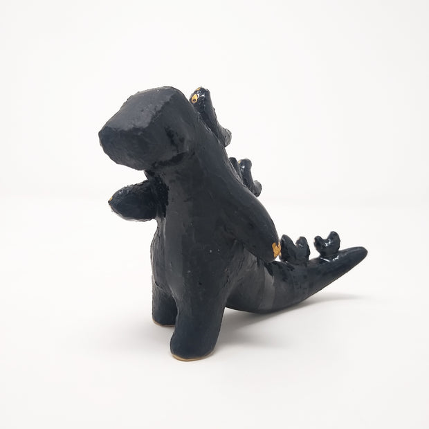 Ceramic sculpture of a dark blue abstract Godzilla figure, with abstract spikes on its back, no facial features, and one arm extended out.