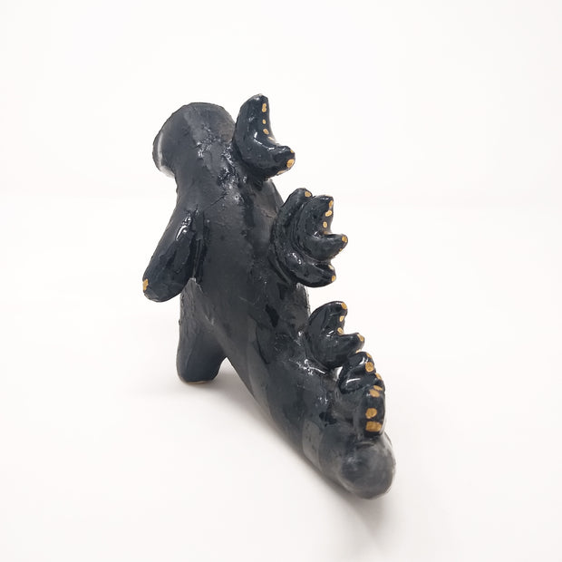 Ceramic sculpture of a dark blue abstract Godzilla figure, with abstract spikes on its back, no facial features, and one arm extended out.