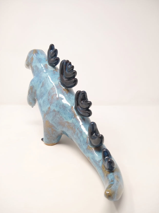 Ceramic sculpture of a brown and blue abstract Godzilla figure, with abstract dark blue spikes on its back, no facial features, and one arm extended out in front.