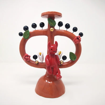Ceramic sculpture of a small red devil sitting on a brown vessel with 2 large handles and a skinny middle. Small leaves and flowers decorate the vessel with black circles protruding out via wire.