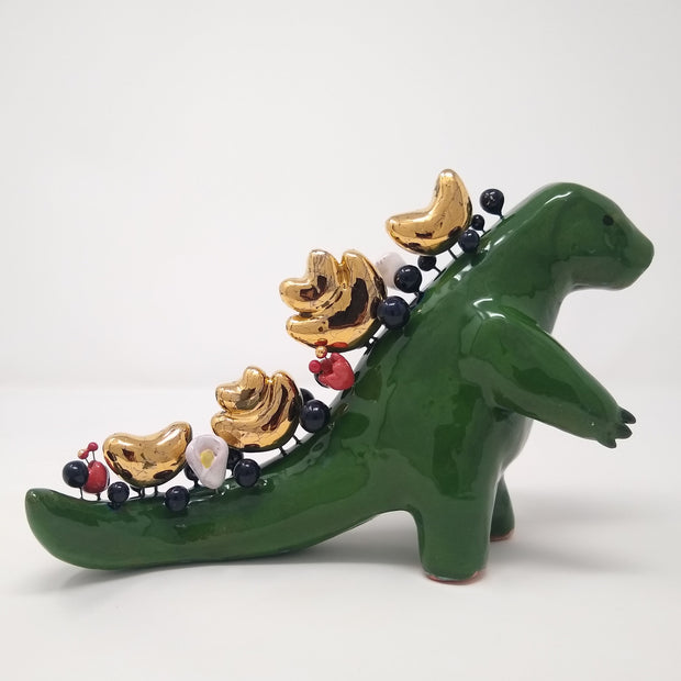 Sculpture of a green Godzilla like monster, standing with its arms slightly out in front of it. Its back and tail are lined with various abstract sculptural shapes and small balls, consisting of gold, black and red and white flowers.