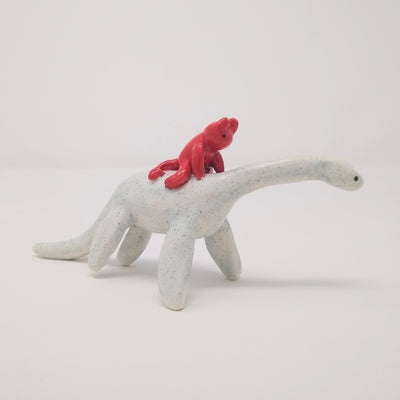 Ceramic sculpture of a small red devil character, simplistic in shape and with a drawn on face, riding atop of a white Plesiosaurus dinosaur. 