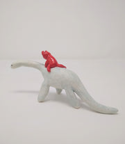 Ceramic sculpture of a small red devil character, simplistic in shape and with a drawn on face, riding atop of a white Plesiosaurus dinosaur.