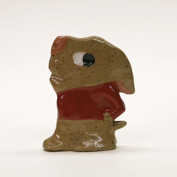 Ceramic sculpture of a flat brown dog with cartoonish large head and eyes that are looking behind him. He has his hand in his pockets and wears a red shirt.