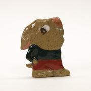 Ceramic sculpture of a flat brown dog with cartoonish large head and eyes that are looking behind him. He has his hand in his pockets and wears a blue shirt with red shorts.