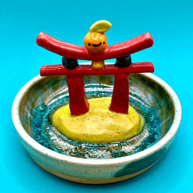 Ceramic sculpture of a red gate with a small orange citrus on top, within a ceramic bowl.