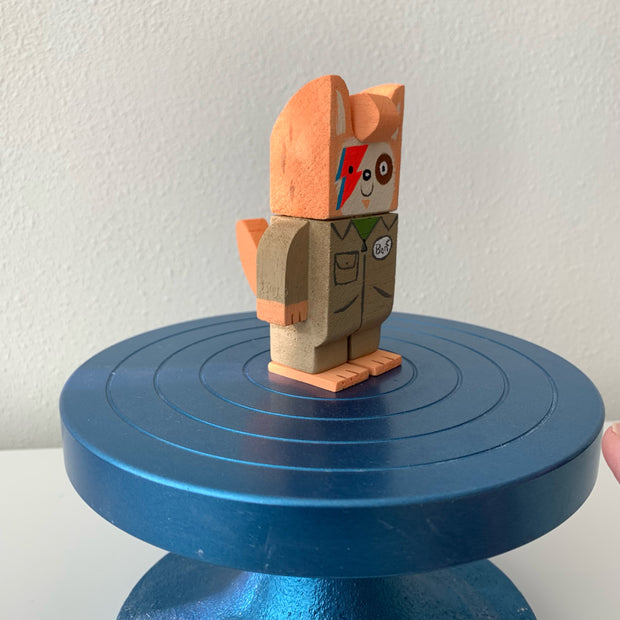 Painted carved wooden sculpture of a dog, with orange coloring and a cream colored face. It has blue and red lightning bolts over one eye, ala David Bowie. It wears a tan jumpsuit that says "Barf" on it. Side view.