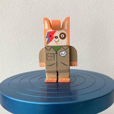 Painted carved wooden sculpture of a dog, with orange coloring and a cream colored face. It has blue and red lightning bolts over one eye, ala David Bowie. It wears a tan jumpsuit that says "Barf" on it.