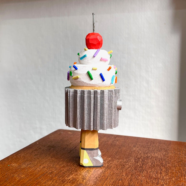 Wooden sculpture of a cupcake with white frosting, sprinkles, and a cherry on top. Its wrapped in a silver wrapper and stands on 2 legs, with a pair of eyes as its only facial feature.