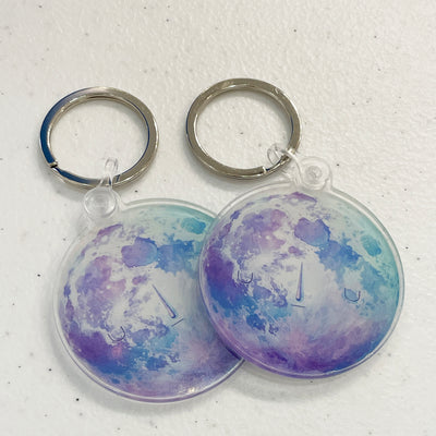 2 round keychains that resemble moons, with blue and purple coloring. They each have small faces that are closed eye and solemn.