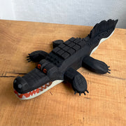Whittled wooden sculpture of an alligator, painted all black and red eyes and a red mouth. Its underside is gray. Its body has a slightly blocky look to it, with its back being a row of cubes.