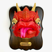 Sculpture of a red cartoonish dragon head with gold horns, gold eyes and a tongue out. Its mounted to a wooden plaque, like a hunter's trophy.