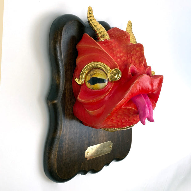 Sculpture of a red cartoonish dragon head with gold horns, gold eyes and a tongue out. Its mounted to a wooden plaque, like a hunter's trophy.
