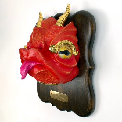 Sculpture of a red cartoonish dragon head with gold horns, gold eyes and a tongue out. Its mounted to a wooden plaque, like a hunter's trophy. 
