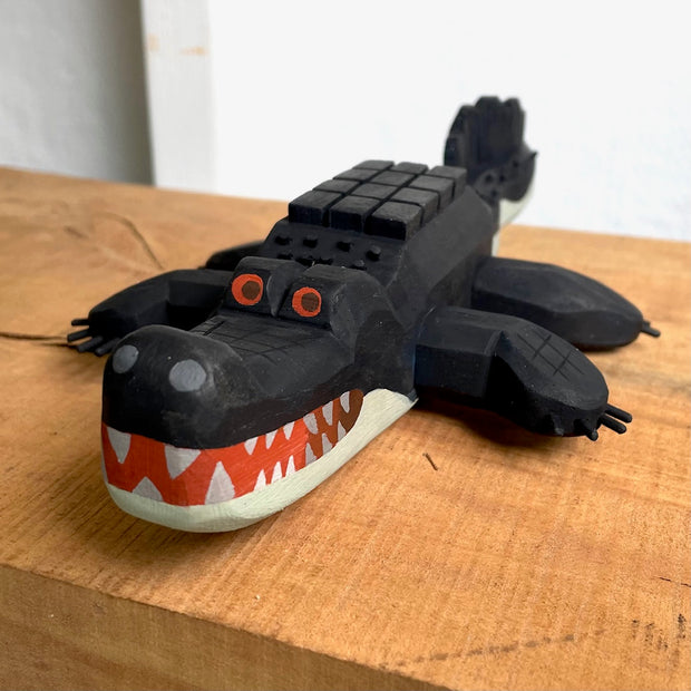 Whittled wooden sculpture of an alligator, painted all black and red eyes and a red mouth. Its underside is gray. Its body has a slightly blocky look to it, with its back being a row of cubes.