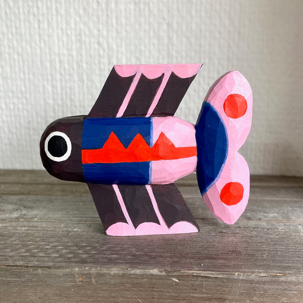 Brightly painted whittled wooden sculpture of a fish colored black with blue and pink body and tail and red accent coloring. It has a set of eyes, one on each side of its head.