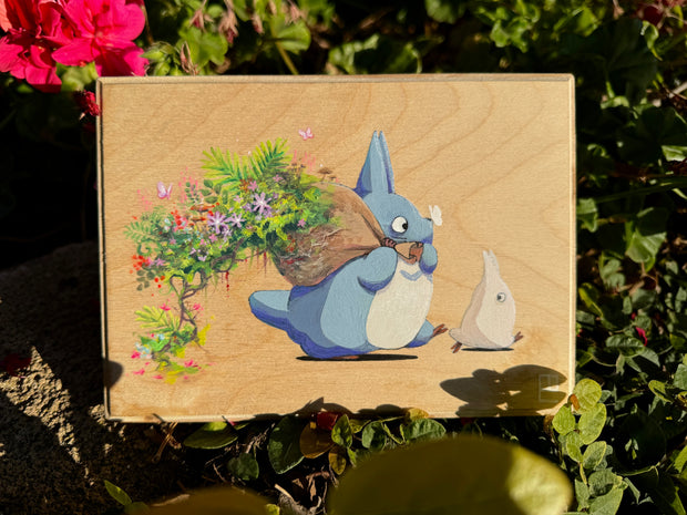 Painting on exposed wooden panel of a blue Chibi Totoro, holding a brown sack over its shoulder. Lush greenery and flowers burst from the bag behind.
