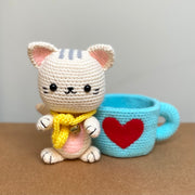 Crochet sculpture of a white cat in a cute, chibi style wearing a yellow scarf and sitting next to a blue mug with a red heart on the exterior.