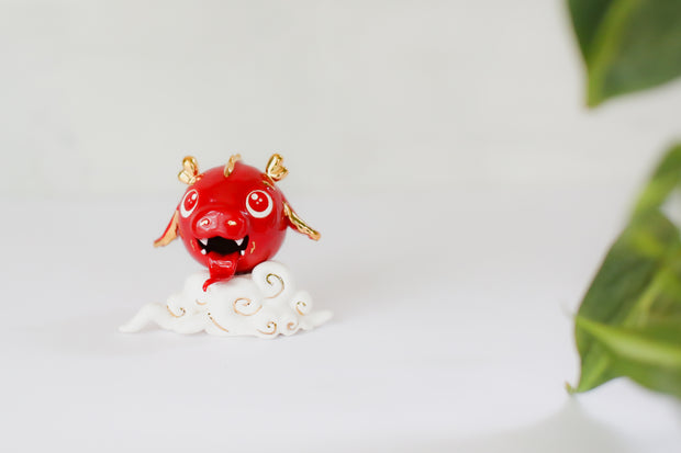 Glazed ceramic sculpture of a cute red dragon head with gold color accents. It's propped up on a cloud and smiles with its tongue out.