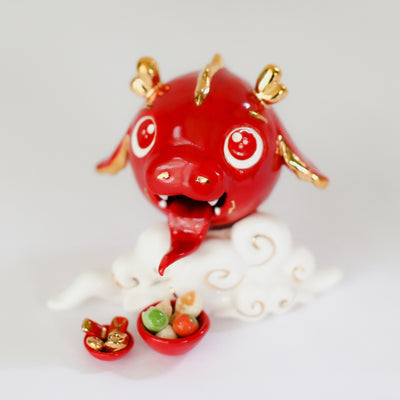 Glazed ceramic sculpture of a cute red dragon head with gold color accents. It's propped up on a cloud and in front are 2 bowls, one with food and the other with gold tokens.