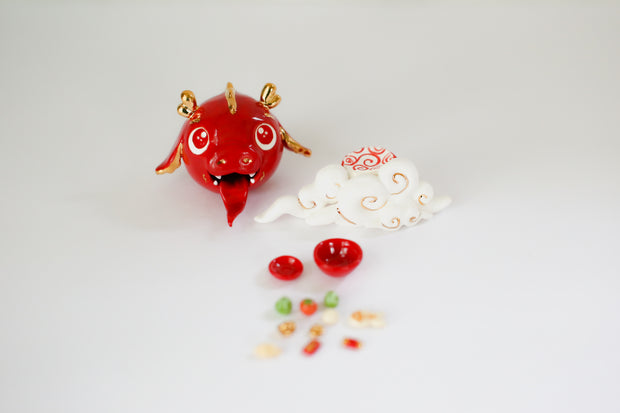 Glazed ceramic sculpture of a cute red dragon head with gold color accents. It's propped up on a cloud and in front are 2 bowls, one with food and the other with gold tokens.