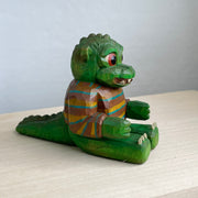 Painted whittled wooden sculpture of a baby Godzilla, with cute large eyes and a smiling sharp toothed expression. It sits on the ground with its arms out in front and wears a brown and blue striped sweater.