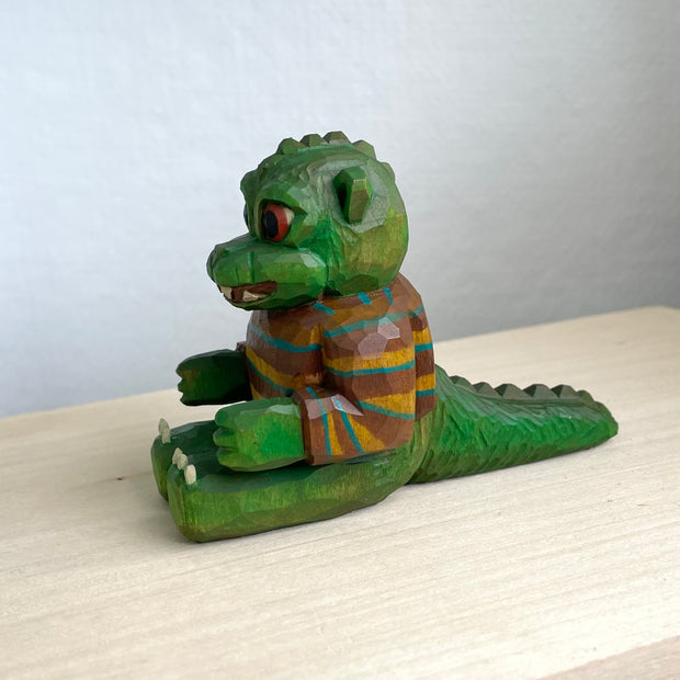 Painted whittled wooden sculpture of a baby Godzilla, with cute large eyes and a smiling sharp toothed expression. It sits on the ground with its arms out in front and wears a brown and blue striped sweater.