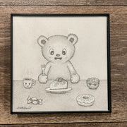 Graphite drawing of a cartoon bear, smiling and sitting at a table with a fork in hand. On the table is a slice of cake, a donut, a mug and candy. Piece is in thin black frame.