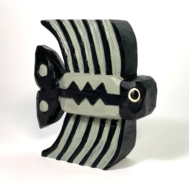 Painted whittled wood sculpture of a fish with large tall fins. The fish is grey and black stripes, with a Charlie Brown esque stripe along its body.