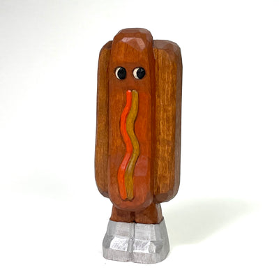 Stained and subtly painted whittled wood sculpture of a hot dog, standing on 2 legs wearing a pair of silver shoes. It looks off to the side and has squiggles of ketchup and mustard.